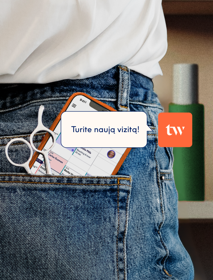 Išmanusis telefonas ir kirpėjo žirklės, įkišti į galinę džinsų kišenę; ekrane matomas Treatwell programėlės rezervacijų kalendorius su pranešimu „naujas vizitas".