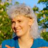 A young woman with a curly white blonde mullet haircut sitting in a park on a sunny day.