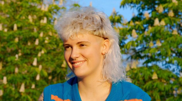 A young woman with a curly white blonde mullet haircut sitting in a park on a sunny day.