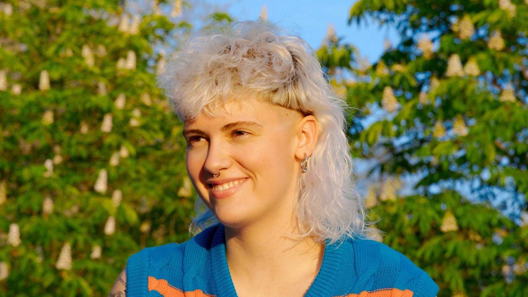 A young woman with a curly white blonde mullet haircut sitting in a park on a sunny day.