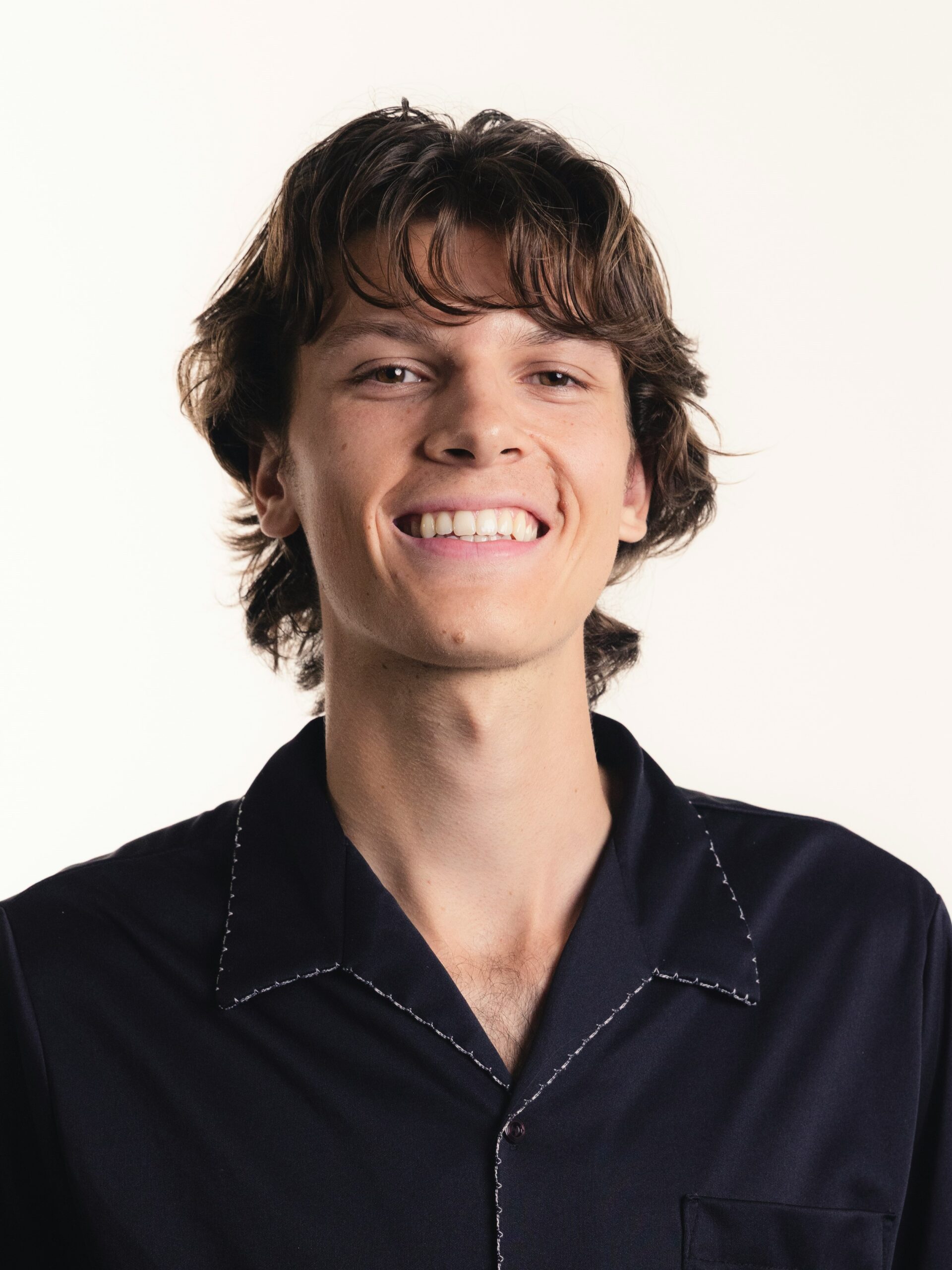 A young man with a layered, textured mullet haircut wearing a dark blue shirt smiling.
