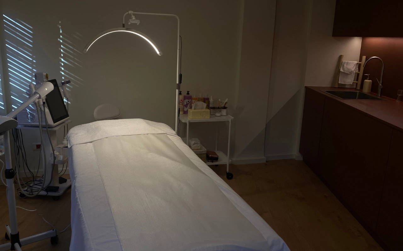 Tiksli procedūrų patalpa su balta lova po lankiniu LED šviesos prietaisu, profesionalia estetikos įranga su skaitmeniniu ekranu, odos priežiūros priemonėmis ant vežimėlio ir tamsaus medžio spintele su kriaukle tolimojoje sienoje.