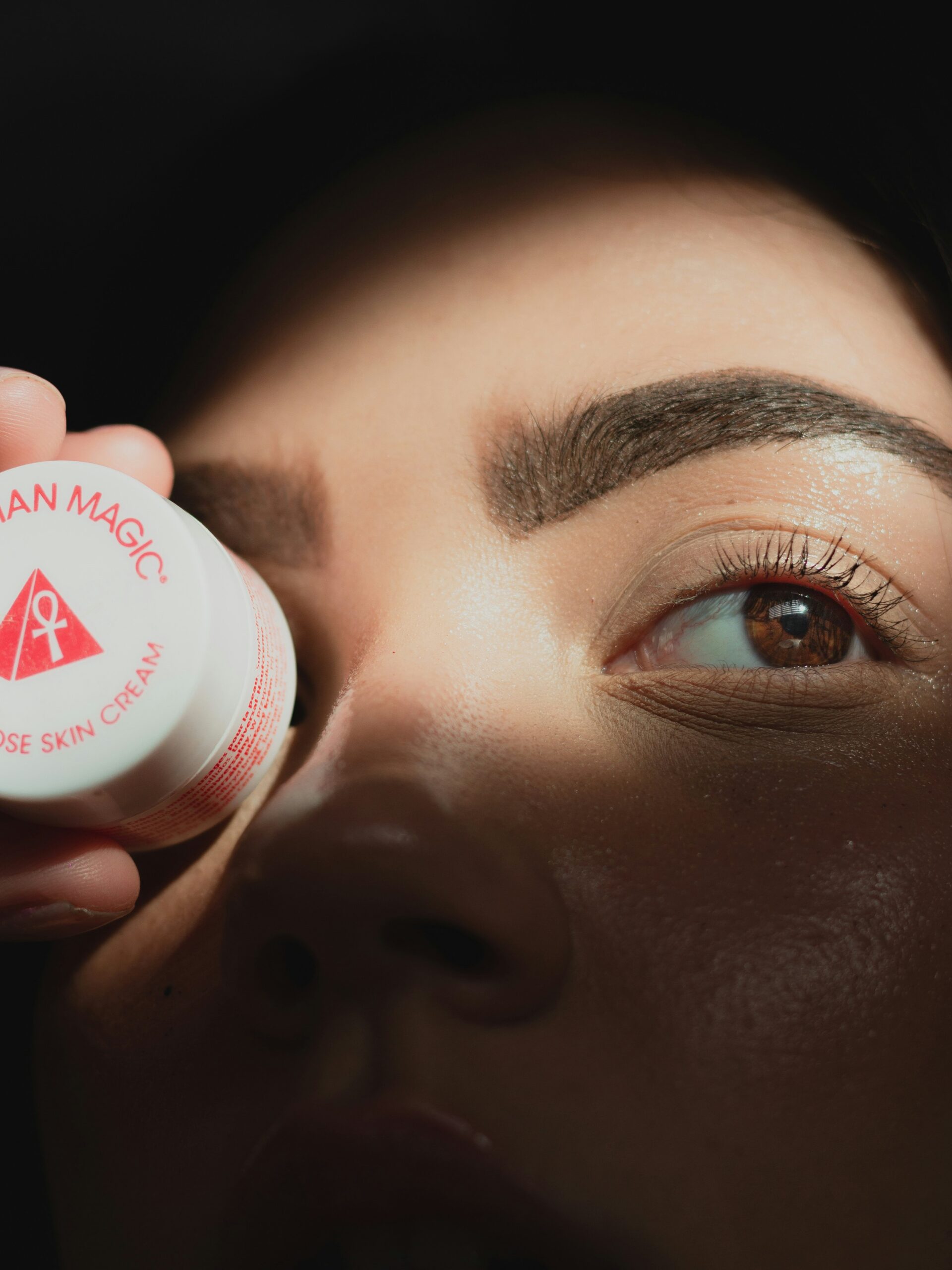 Moters veidas su microblading antakiais iš arti, laiko mažą apvalų kremo indelį priešais vieną akį