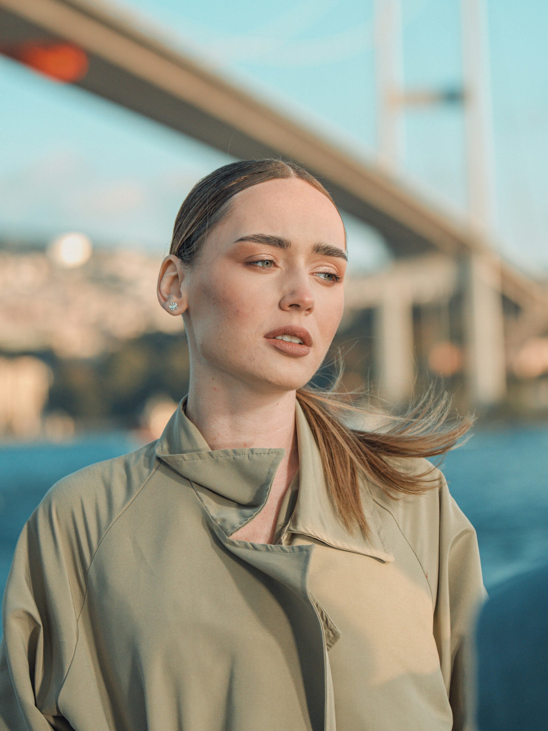 Moteris su microblading antakiais ir surištais šviesiais plaukais, vilkinti lietpaltį, stovi priešais tiltą