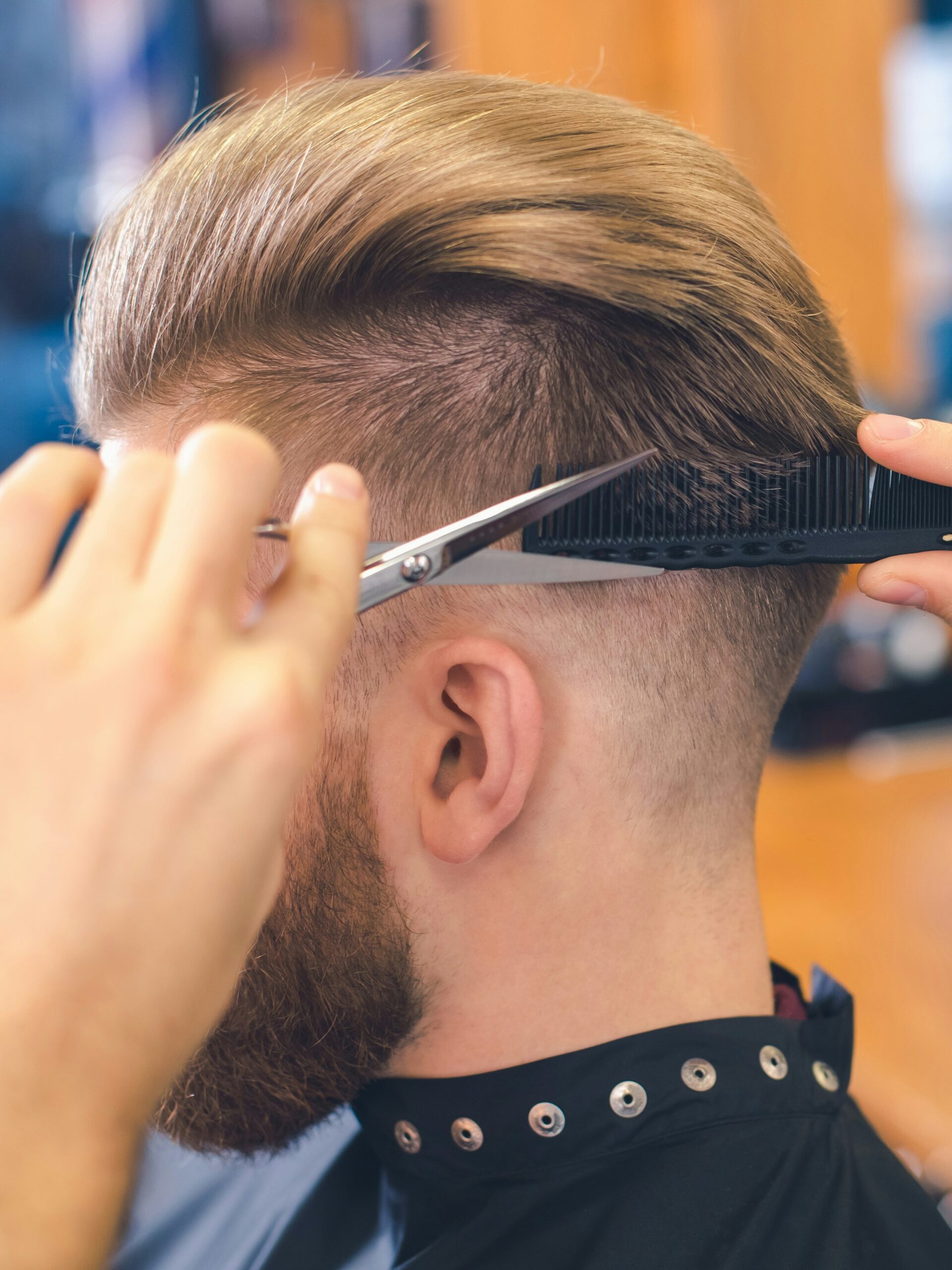 Artimas vaizdas: barberis kirpdamas taper fade klientui naudoja žirklių virš šukų techniką pakaušio srityje