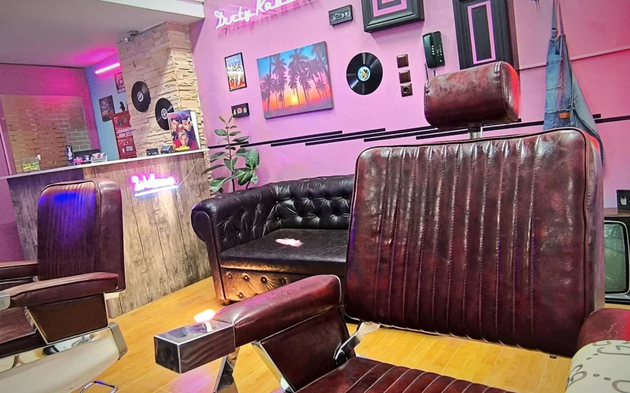 Retro stiliaus barberainė su rožinėmis ir violetinėmis sienomis, neoniniu užrašu, vinilo plokštelėmis kaip dekoru, palmių saulėlydžio tapyba, tamsios odos barberių kėdėmis ir Chesterfield sofa ant šiltų medinių grindų.