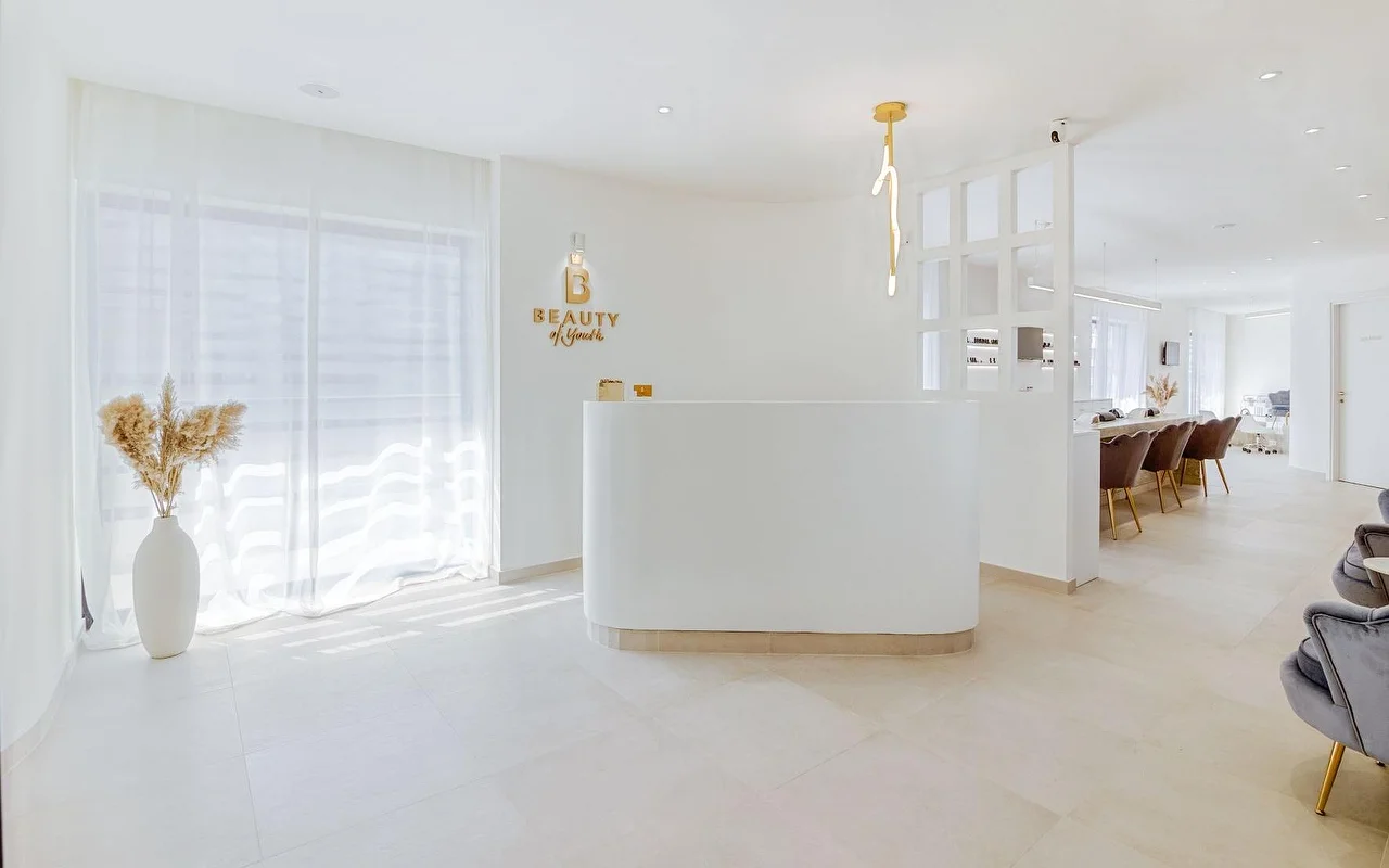 Šviesus, visiškai baltas priėmimo zona su lenktu registracijos stalu, auksiniu Beauty of Youth logotipu ant sienos, skulptūriniu pakabučiu, natūralią šviesą filtruojančiomis užuolaidomis ir rudos aksomo kėdėmis fone.