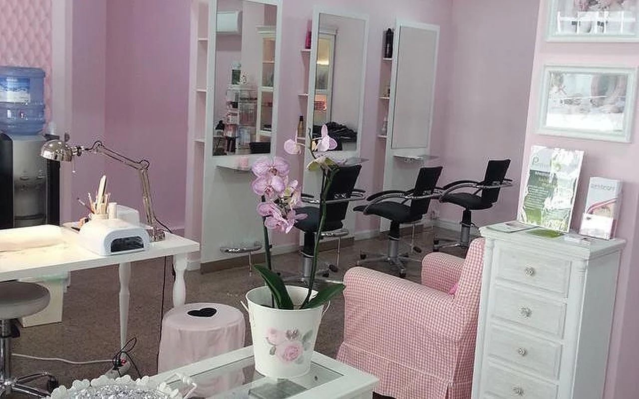 Ryškaus, rožinių atspalvių plaukų ir nagų salono interjeras. Kairėje – balta manikiūro stotelė su staline lempa ir nagų kietinimo aparatu. Centre – orchidėja baltame vazone ant kavos staliuko šalia rožinės ir baltos languotos kėdės. Fone – kelios plaukų kirpimo vietos su aukštais veidrodžiais ir juodomis saloninėmis kėdėmis prie šviesiai rožinių sienų.