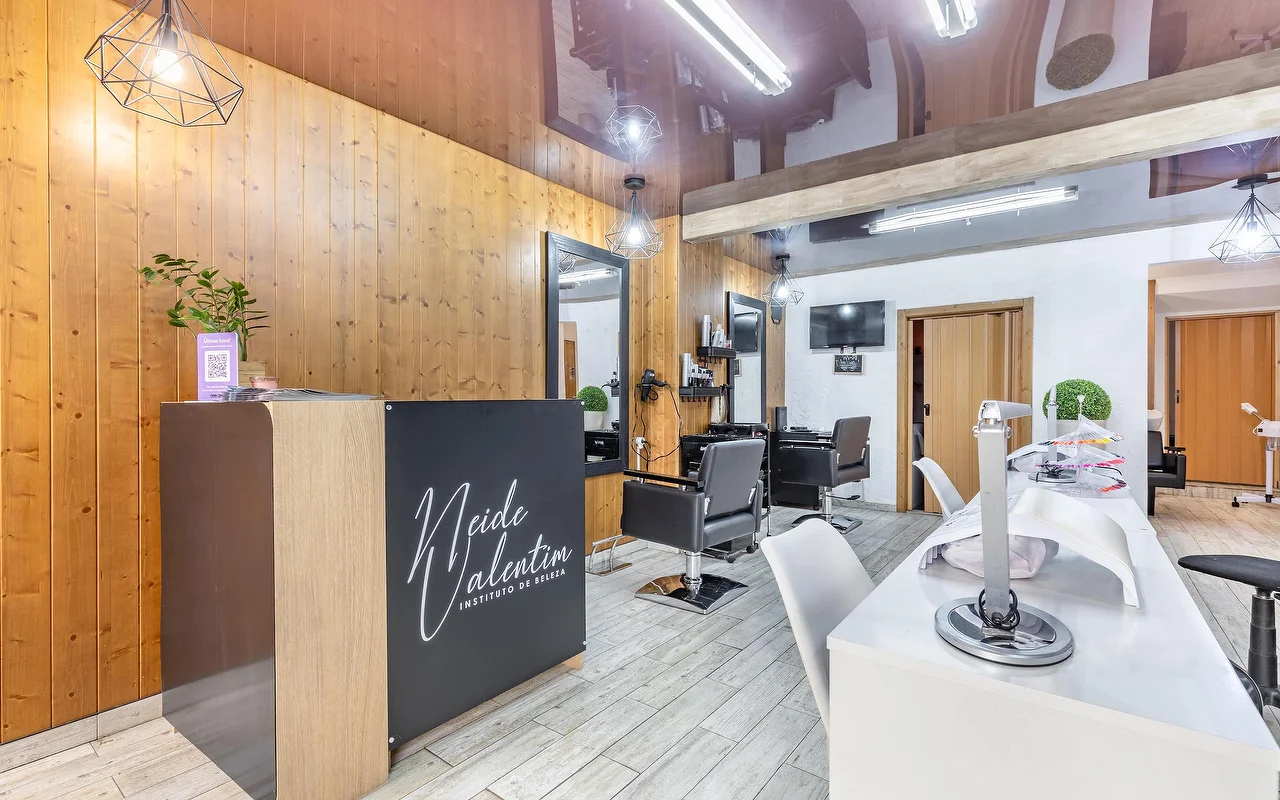 Modernus grožio salonas su registratūros stalu kairėje, pažymėtu Neide Valentim Instituto de Beleza. Sienos apdailintos šiltomis vertikaliomis medinėmis plokštėmis ir dideliais veidrodžiais. Priešais veidrodžius stovi juodos stilistų kėdės, o dešinėje – baltos manikiūro stotelės su baltomis kėdėmis ant pilkų plytelių grindų.
