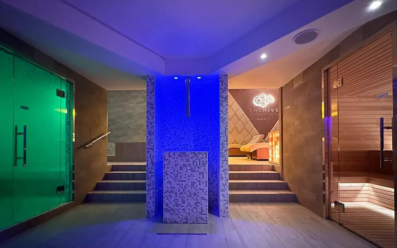Prabangaus SPA terminė zona su dramatiška mėlyna ir žalia atmosferine apšvietimu. Erdvėje — medinė suomiška pirtis, garų pirties įėjimas ir centrinė mozaikinė konstrukcija su laiptais, vedančiais į poilsio zonas.