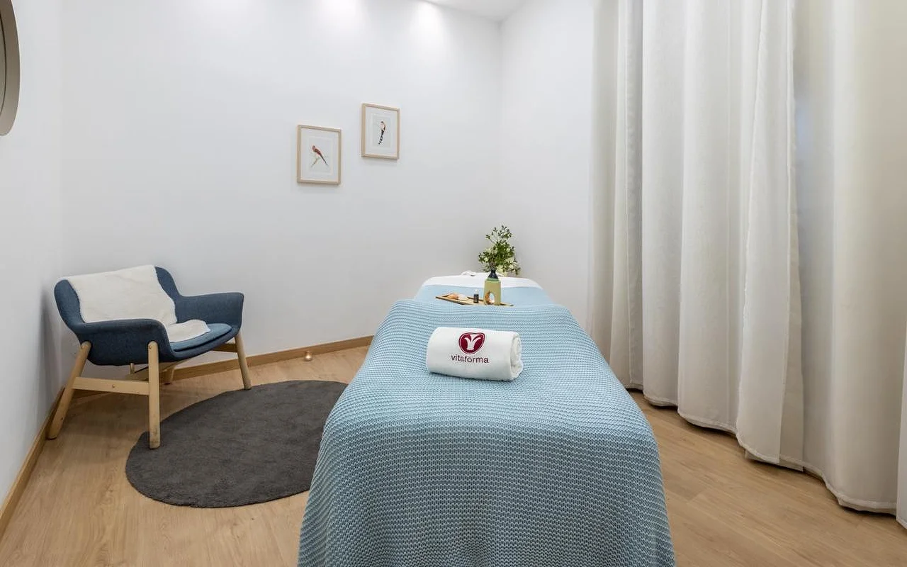 Švari, minimalistinė SPA procedūrų kabina su baltomis sienomis ir šviesiomis medinėmis grindimis. Procedūrinis stalas užtiestas šviesiai mėlyna megzta antklode ir suvyniotas baltas rankšluostis su Vitaforma logotipu. Kampe – stilingas mėlynas fotelis ant mažo apvalaus pilko kilimėlio, o prie lango kabo ilgos baltos užuolaidos.