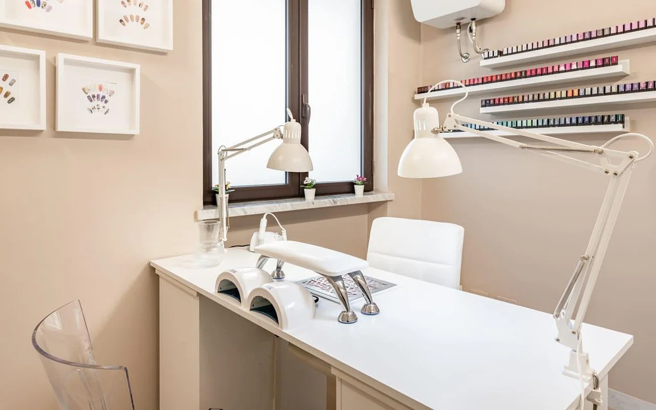 Nagų stotelė AG The Beauty Room's salone su &scaron;variu baltu stalu, dviem UV lempomis, reguliuojamomis darbo lempomis ir lentynomis, kuriose rikiuojasi eilės spalvotų nagų lakų. Ant &scaron;iltų atspalvių sienų kabo įrėminti nagų dizaino darbai.
