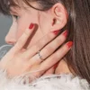 Brunetės moters galvos profilis i&scaron; arti, ranka liečia veidą, matosi blizgus raudonas gelinis manikiūras