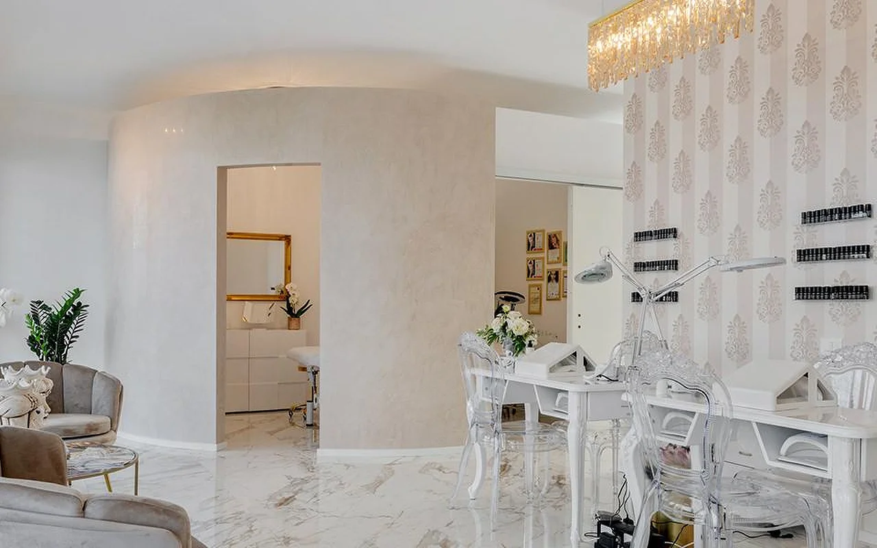 Eleganti&scaron;kas MC Beauty Center interjeras su marmuro imitacijos grindimis, baltomis i&scaron;lenktomis sienomis ir auksiniu kri&scaron;tolo sietynu. De&scaron;inėje – nagų stotelė su skaidriomis kėdėmis, o pro auksu įrėmintas duris matyti procedūrų kabinetas su aksominiais foteliais.