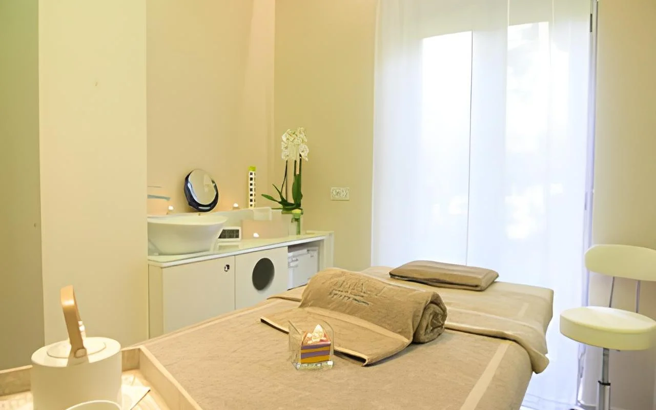 Ramus procedūrų kabinetas Alice Beauty Clinique su ku&scaron;ete, paklota mink&scaron;ta pilk&scaron;vai rusva patalyne, baltomis orchidėjomis ant stalvir&scaron;io, žvakėmis prie praustuvo ir permatomomis užuolaidomis, filtruojančiomis natūralią &scaron;viesą pro didelį langą.