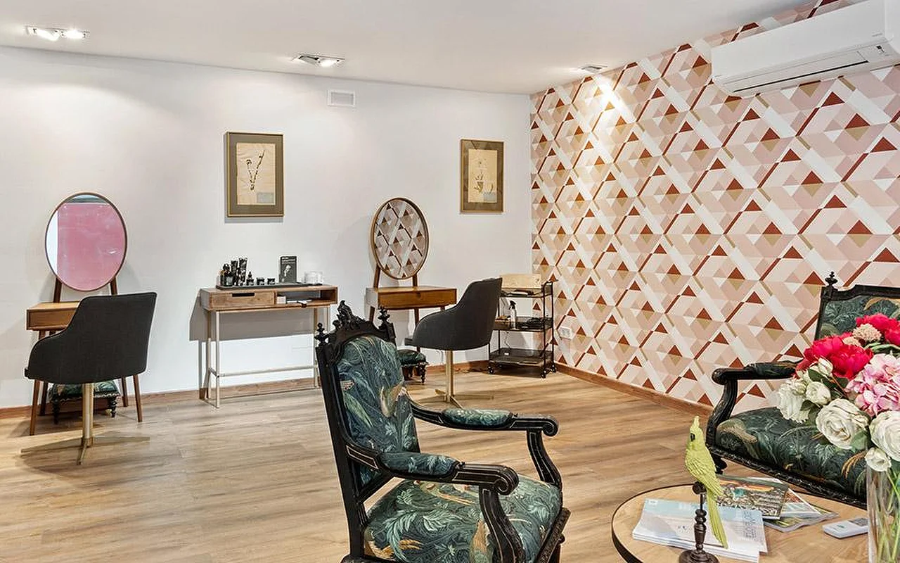 Elegantiškas salono kambarys su geometriška rožinės ir terakotos spalvos akcentine siena, pora puošnių drožinėtų kėdžių, aptrauktų žaliu augaliniu raštu audiniu, mediniai stilizavimo staliukai su apvaliais veidrodžiais ir rožinių bei raudonų gėlių vaza priekiniame plane.