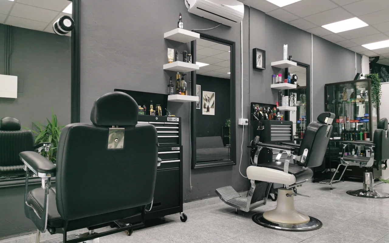 Tradicinis barberio salonas su angliškai pilkomis sienomis, klasikinėmis juodos odos barberio kėdėmis su chromuotais pagrindais ir pakojomis, baltomis kabančiomis lentynomis su priežiūros priemonėmis ir dideliais veidrodžiais ant plytelėmis išklotų grindų.