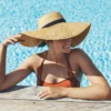 Moteris saulėtame lauko baseine, atsirėmusi į kra&scaron;tą, su oranžiniu bikiniu ir plačiabryle &scaron;iaudine skrybėle