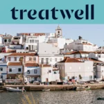 Baltai tinkuotas Algarve žvejų kaimelis su terakotinių stogų namais, kylančiais &scaron;laitu vir&scaron; ramaus vandens, mažos žvejų valtys, priri&scaron;tos palei krantinę, palmės ir bažnyčios bok&scaron;tas po giedru mėlynu dangumi.