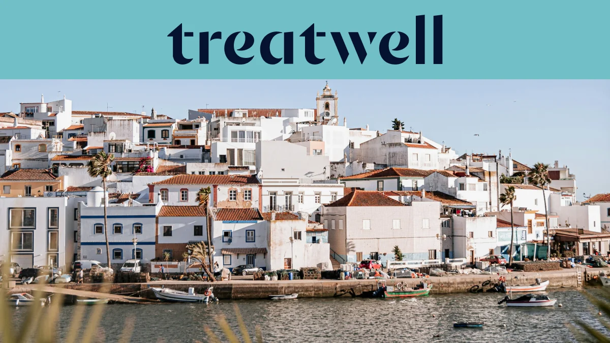 Baltai tinkuotas Algarve žvejų kaimelis su terakotinių stogų namais, kylančiais šlaitu virš ramaus vandens, mažos žvejų valtys, pririštos palei krantinę, palmės ir bažnyčios bokštas po giedru mėlynu dangumi.