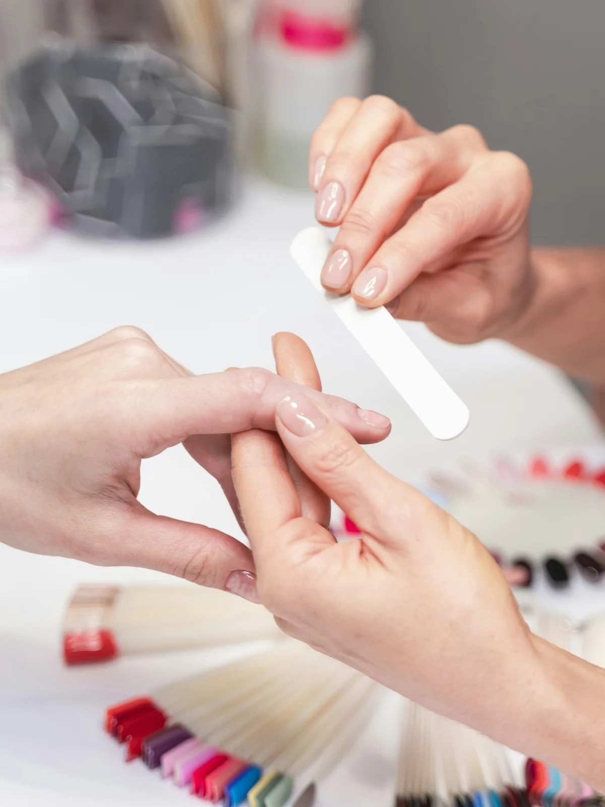 Iš arti — manikiūrininkė švelniai dildo klientės nagus salone, fone matyti manikiūro produktai ir įrankiai