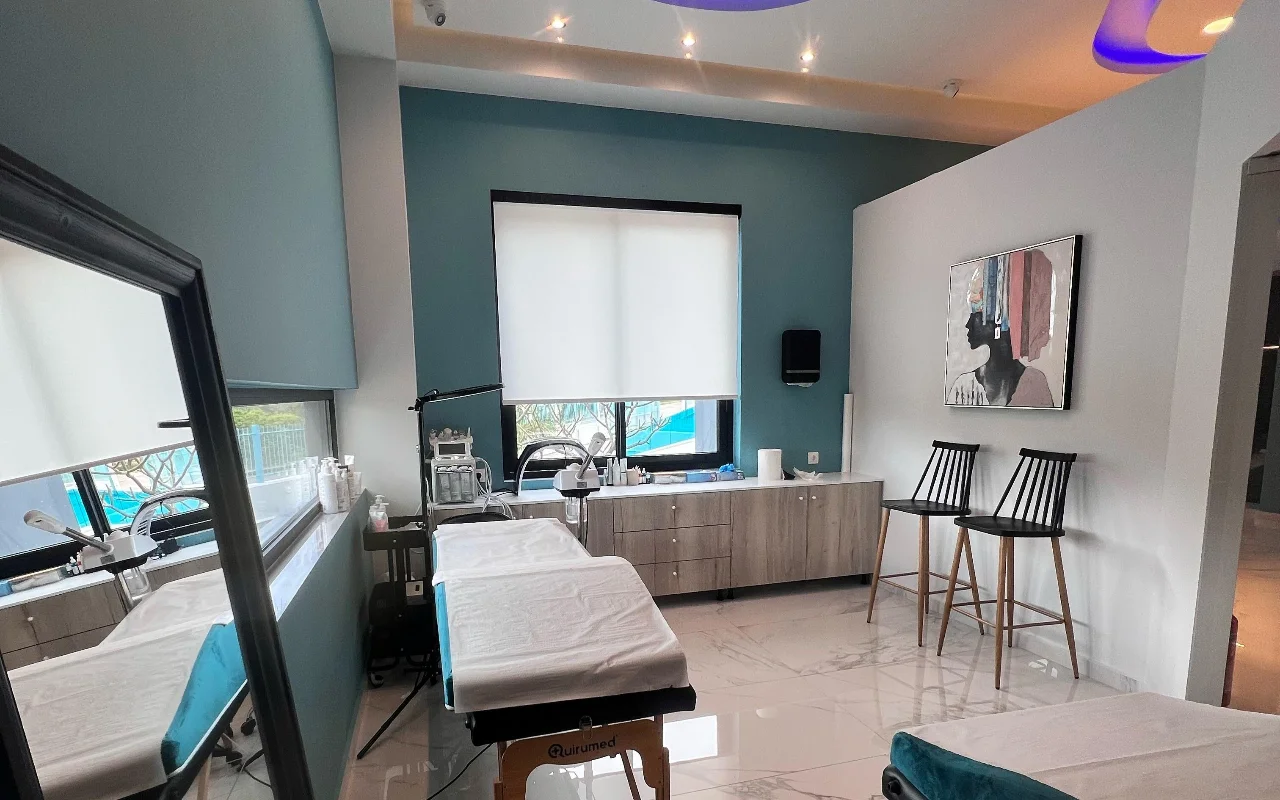 Veido procedūrų patalpa su naftos žalumo sienomis, balta procedūrų kušete, skincare įranga, dideliu veidrodžiu, dviem juodomis lentelėtomis taburetėmis ir įrėmintu portretu, apšviesta lango su baltu roletu.