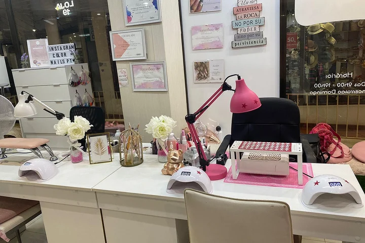 Nagų salono manikiūro stotelė su baltais stalais, ryškiai rausva lempa ir LED lempomis, baltomis rožėmis ir įrėmintais pažymėjimais ant sienos, su pavadinimu Color Nails PT, matomu vitrinoje šalia darbo laiko.
