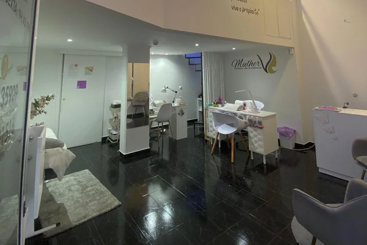 Grožio centro interjeras: tamsios blizgios plytelės ant grindų, balta manikiūro stotelė ir minkštai apšviesti procedūrų plotai, Mulher logotipas ant galinės sienos ir veido procedūros krėslas, matomas už stiklinės pertvaros.