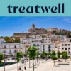 Saulėta panorama į Ibiza miestą, kur baltai i&scaron;balinti pastatai kyla iki katedros ir senųjų Dalt Vila įtvirtinimo sienų ant kalvos. Medžiais apsodinta uosto promenada eina pirmame plane po skaidriai mėlynu dangumi.
