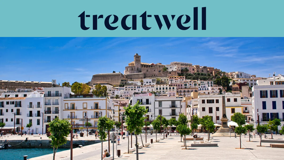Saulėta panorama į Ibiza miestą, kur baltai išbalinti pastatai kyla iki katedros ir senųjų Dalt Vila įtvirtinimo sienų ant kalvos. Medžiais apsodinta uosto promenada eina pirmame plane po skaidriai mėlynu dangumi.
