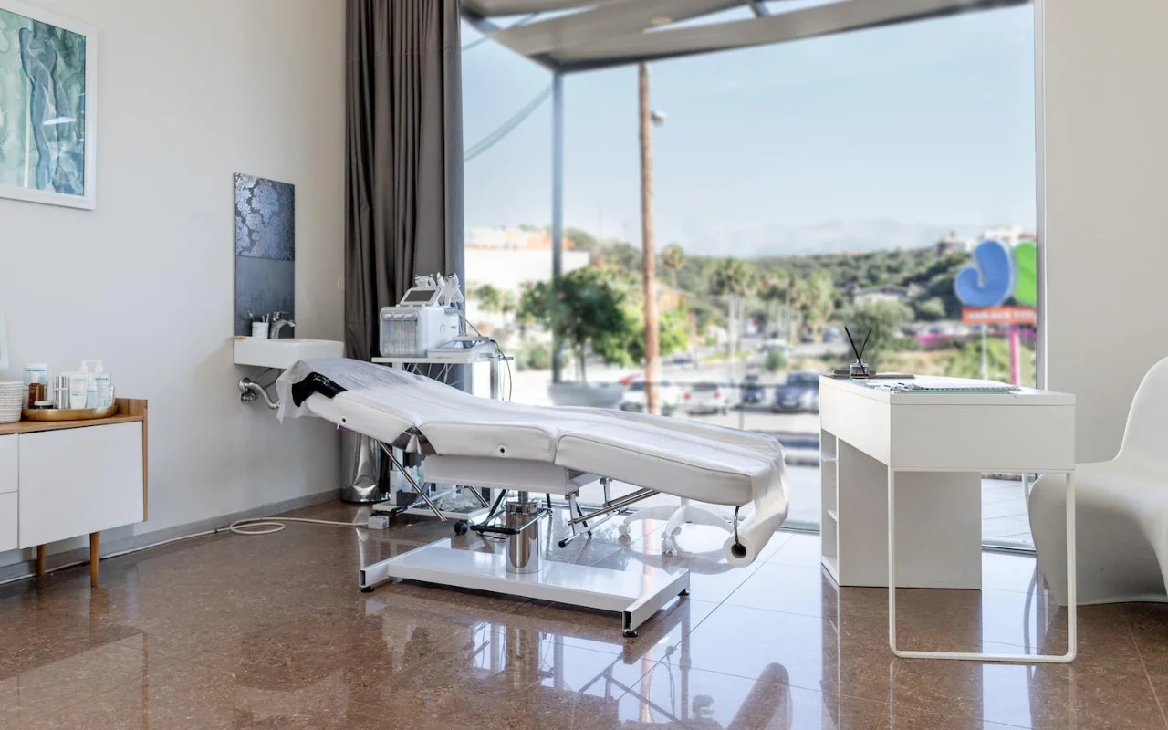 Švari balta procedūrų kabineto erdvė su reguliuojama grožio kušete, skincare įranga ant vežimėlio, įrėmintais meno kūriniais ir dideliu langu, per kurį matyti medžiai, kelias ir tolimos kalvos.