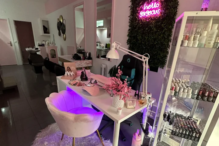 Rausvai apšviestas grožio kampelis su 'hello gorgeous' neoniniu užrašu ant žalios sienos, kreminės spalvos kriauklės formos krėslas, baltas stalas su teptukais ir vyšnių žiedais, ir vitrina, pilna spalvingų nagų lakų.