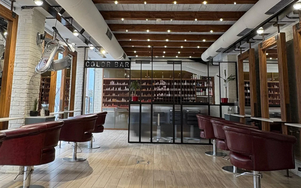 Kirpykla su atviromis medinėmis stogo sijomis ir kalkintomis plytomis, bordo odos styling kėdėmis ir stiklinių sienų dažymo kambariu, apjuostu produktų lentynomis už „Color Bar“ ženklo.