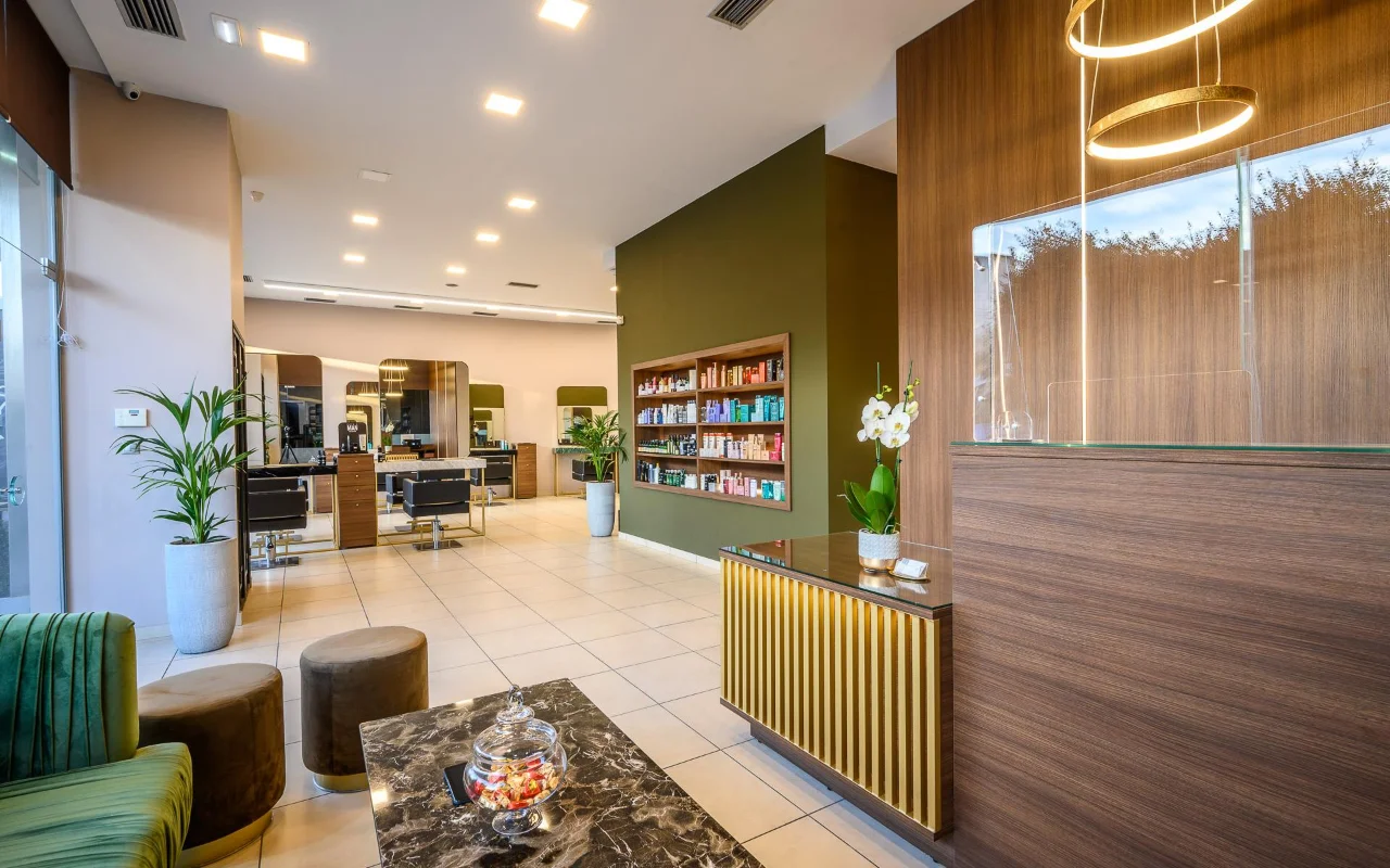 Aukšto lygio kirpykla su šiltais medžio apmušalais, alyvuogių žalio produktų lentynų akcento siena, styling vietomis su dideliais veidrodžiais, registratūros stalu su auksinėmis juostomis ir žalio aksomo sėdynėmis po apvaliais auksiniais pakabinamais šviestuvais.