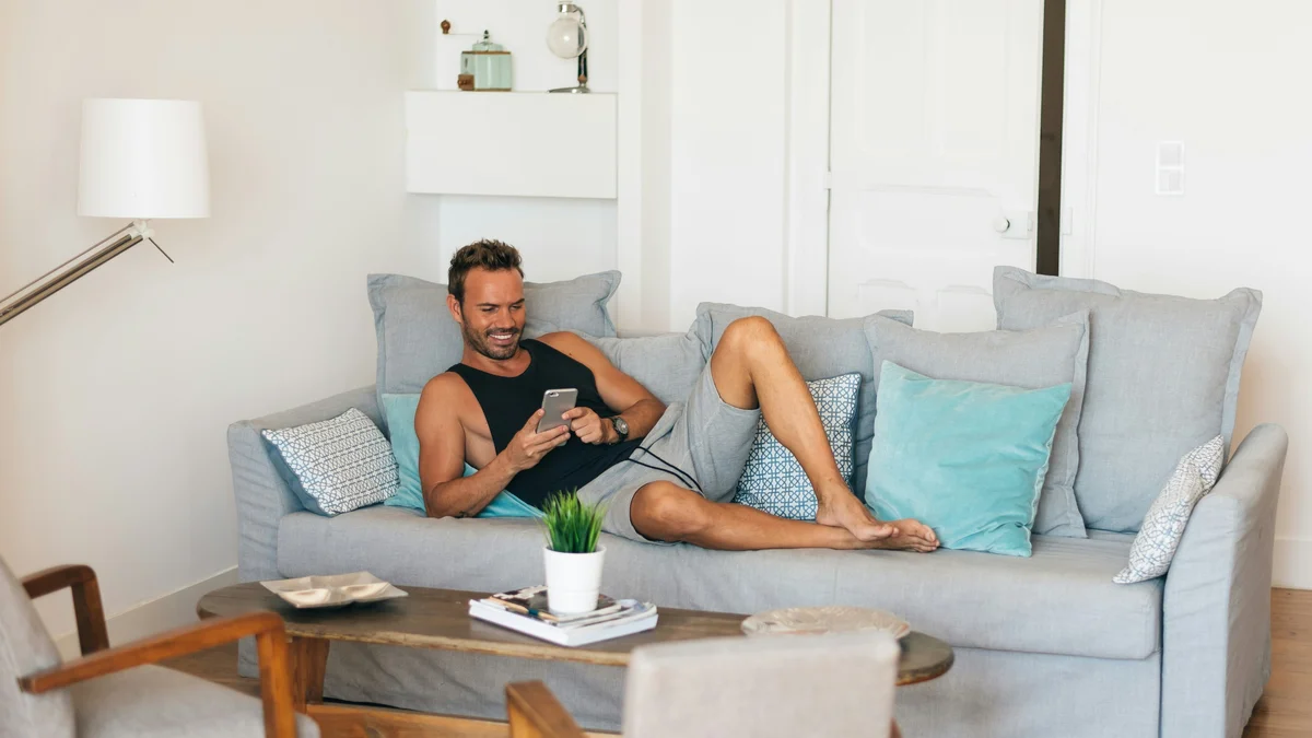 Vyras atsipalaiduoja ant sofos, pakėlęs kojas, ir šypsosi žiūrėdamas į telefoną; jis vilki juodus marškinėlius be rankovių ir laisvus, minkštus pilkus šortus