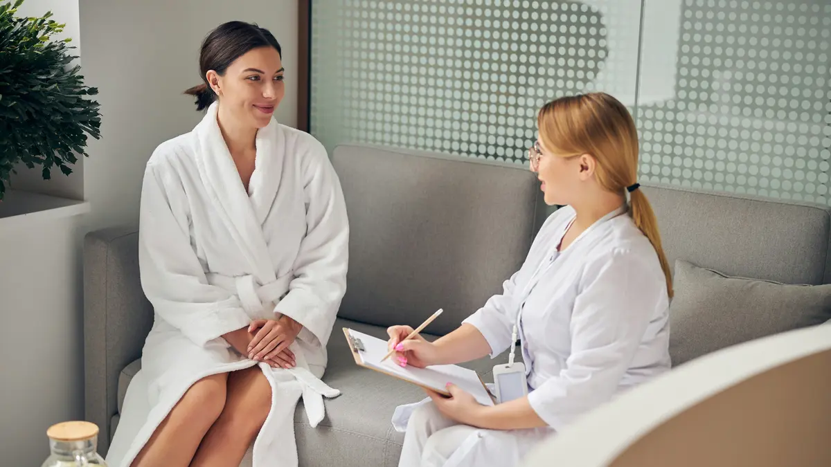 Klientė su baltu chalatu ir specialistė su segtuvu sėdi greta viena kitos ant sofos grožio salone, specialistė užduoda klausimus prieš procedūrą ir juos užsirašo